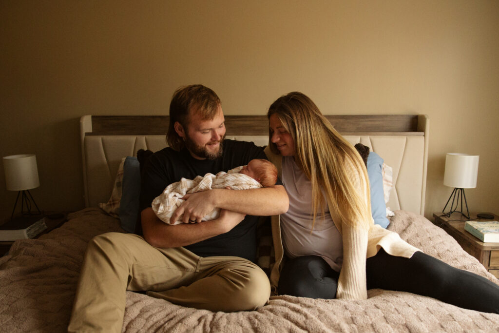 mother and father sitting on a bed holding their baby during newborn photos in traverse city michigan