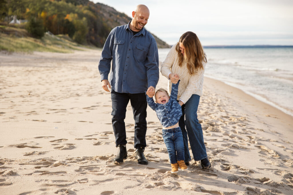 mom and dad swinging little boy on empire beach during fall family portraits in Empire. Photo by Lina Lavonne photography a traverse city photographer