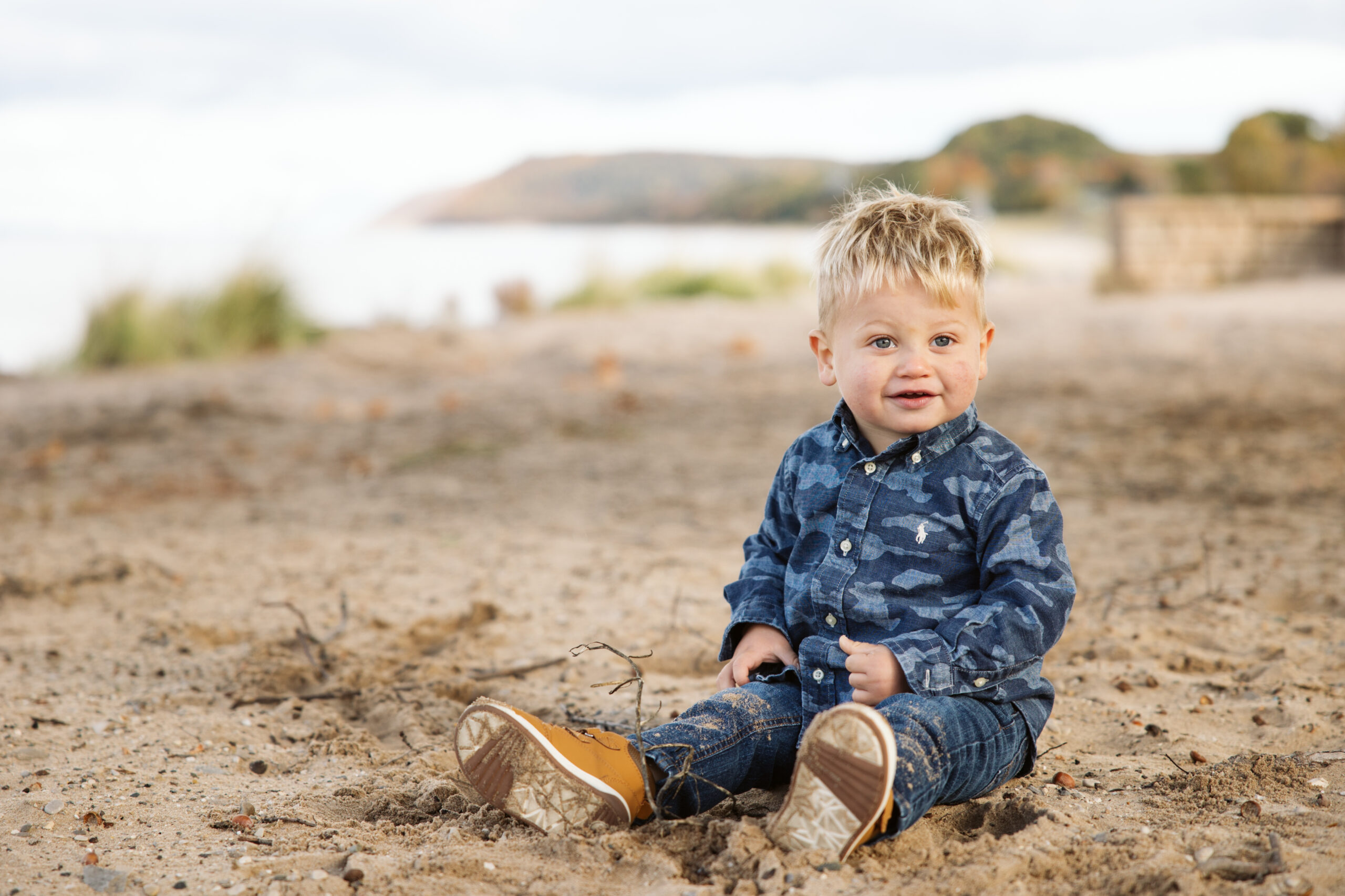 Little boy smiling and sitting on beach during family fall photos. Photo by Lina Lavonne photography a traverse city photographer