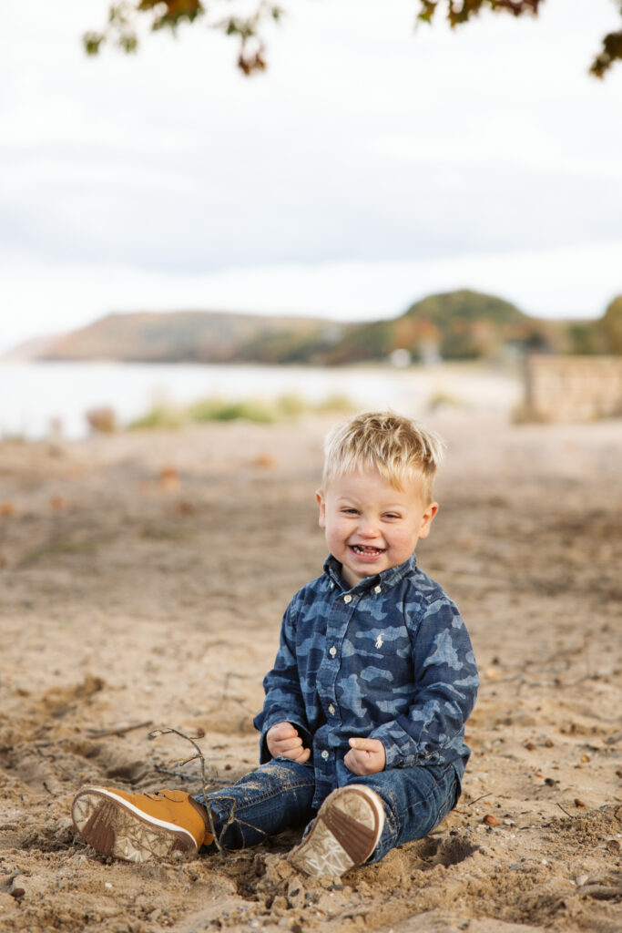 Little boy laughing and sitting on beach during family fall photos. Photo by Lina Lavonne photography a traverse city photographer
