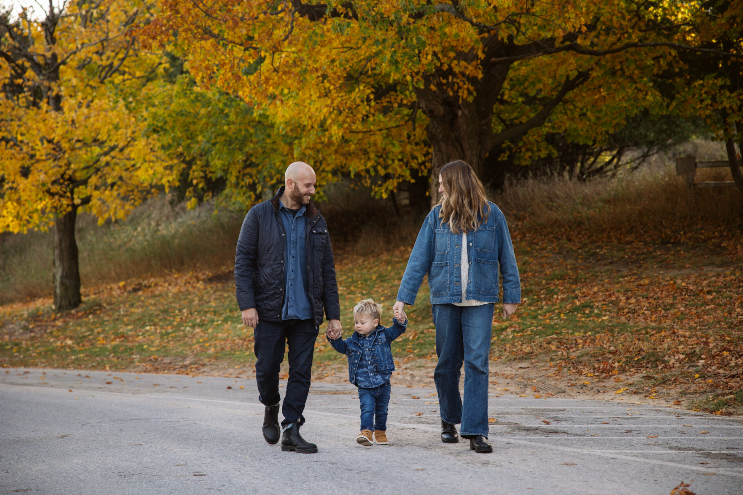 family of three walking down a fall road in northern michigan holding hands during family portraits in Empire. Photo by Lina Lavonne photography a traverse city photographer