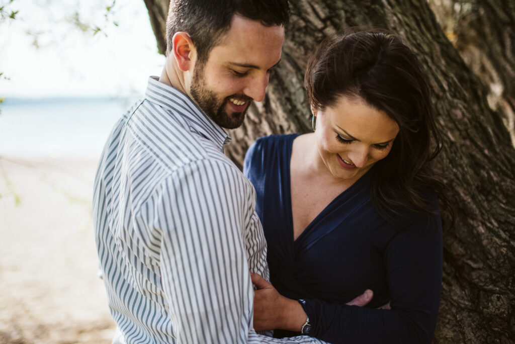 Traverse-City-Michigan-Wedding-Photographer-3 engaged couple hugging against tree on Traverse City beach on Old Mission Peninsula during engagement photos. Photo by Traverse City Photographer, Lina Lavonne Photography