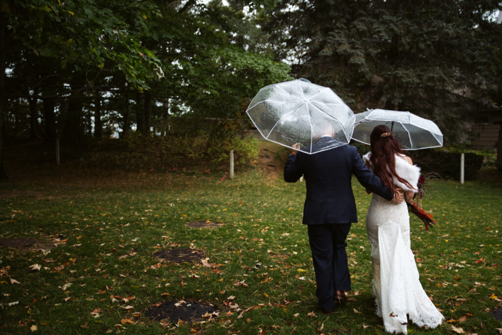 bride and groom walking through a rainy field with umbrellas at peninsula cellars in traverse city